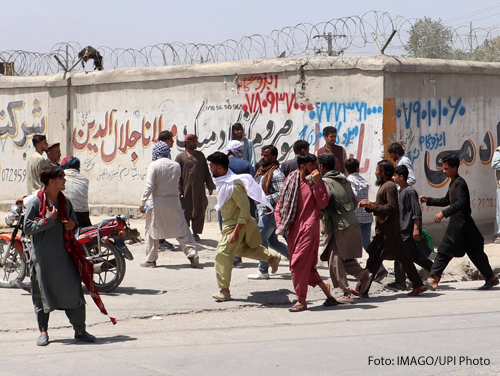 Menschen rennen auf den Flughafen in Kabul zu. Im Hintergrund ist eine Mauer mit Stacheldrahtzaun zu sehen. 