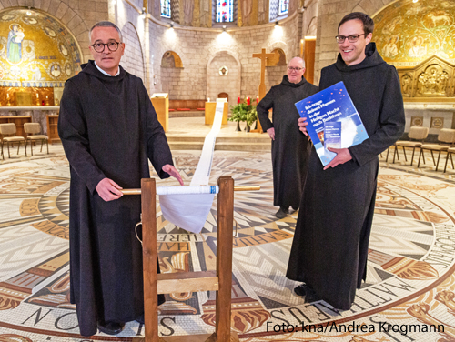 Drei Mönche zeigen eine lange Papierrolle, auf der Gebetsanliegen stehen. 