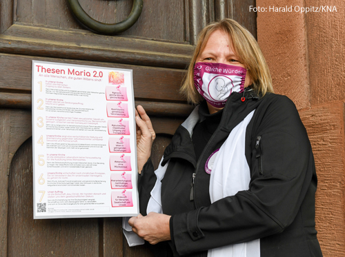 Andrea Keber hält ein Plakat in der Hand, dass sie gleich an eine Kirchentür schlagen wird. Darauf stehen sieben Thesen der Initiative Maria 2.0  für Reformen in der katholischen Kirche. 