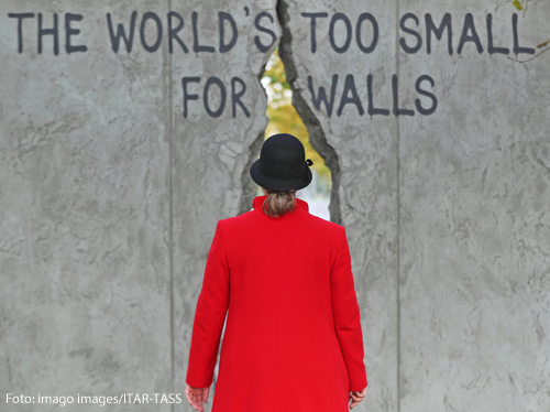 Eine Frau geht durch den Riss einer Mauer. Sie trägt einen schwarzen Hut und einen roten Mantel. 