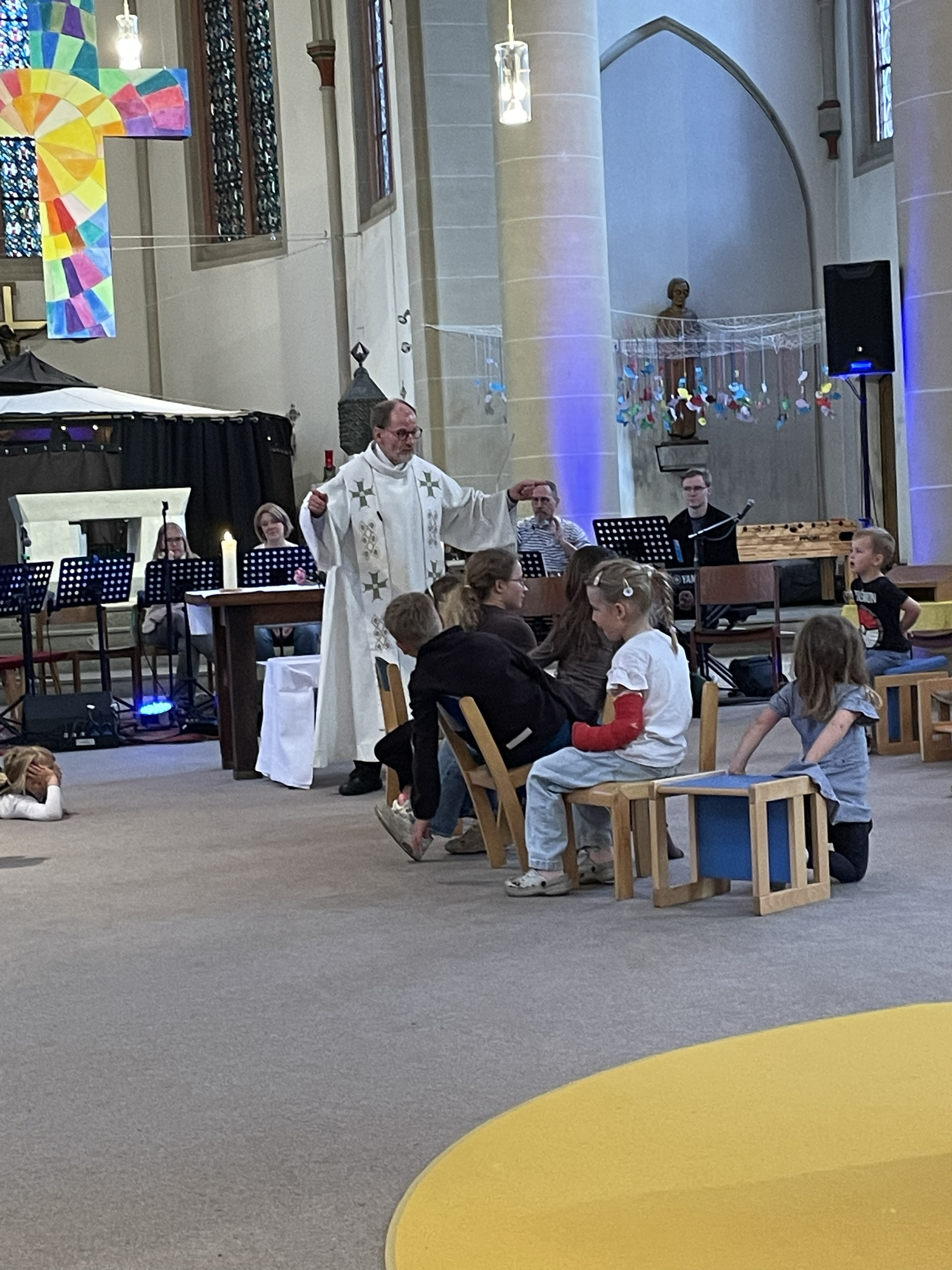 Kinder auf Holzstühlen in einer Kirche, ein Pastor steht davor, im Hintergrund Personen hinter schwarzen Notenständern