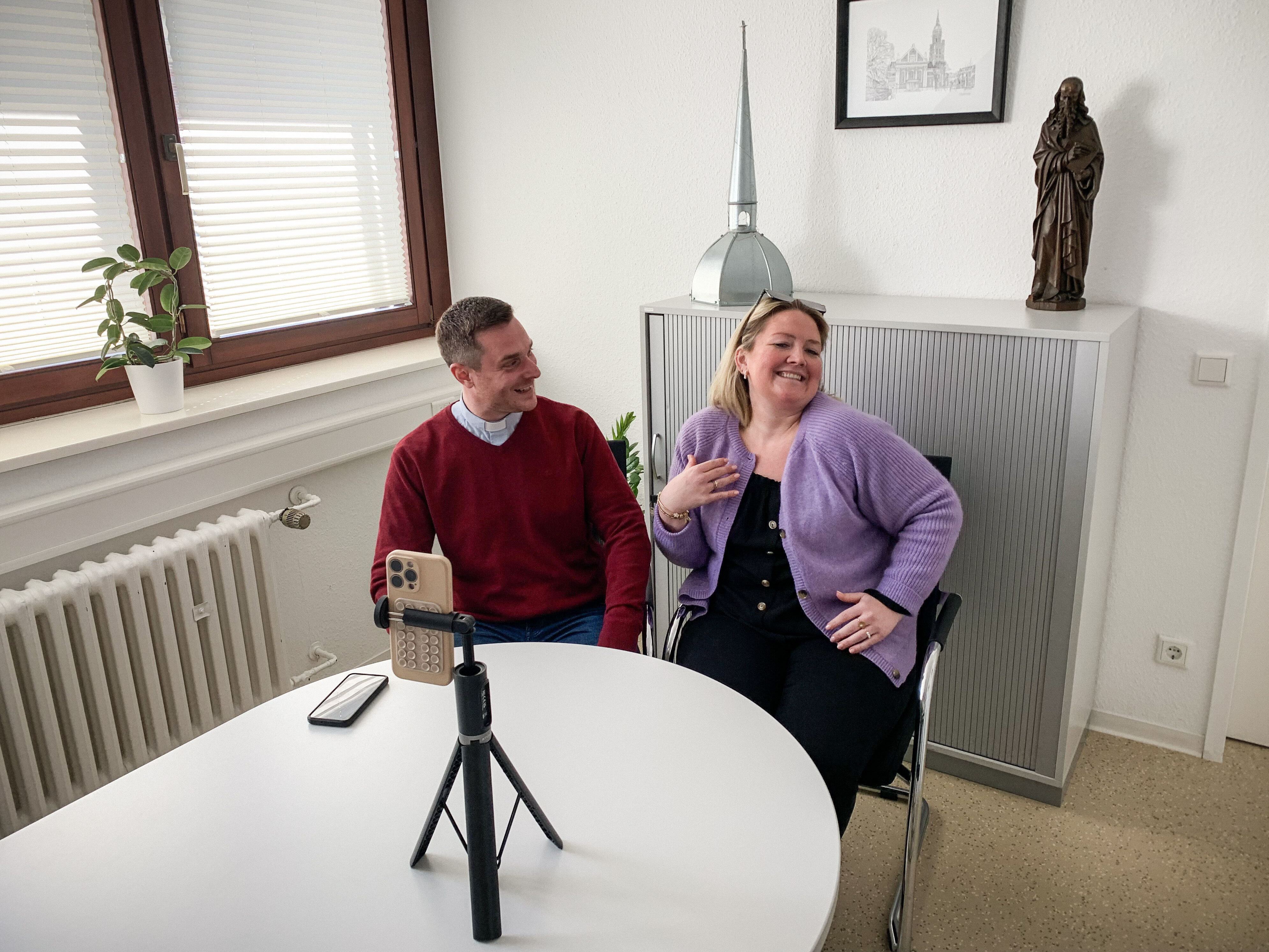 Ein Mann und eine Frau sitzen an einem runden Tisch, schauen in die Kamera eines Smartphones. Das steht in einem kleinen Stativ auf dem Tisch. Hinter ihnen auf einem Schrank steht eine Marienstatue und das Dach eines Kirchturms als Requisiten.