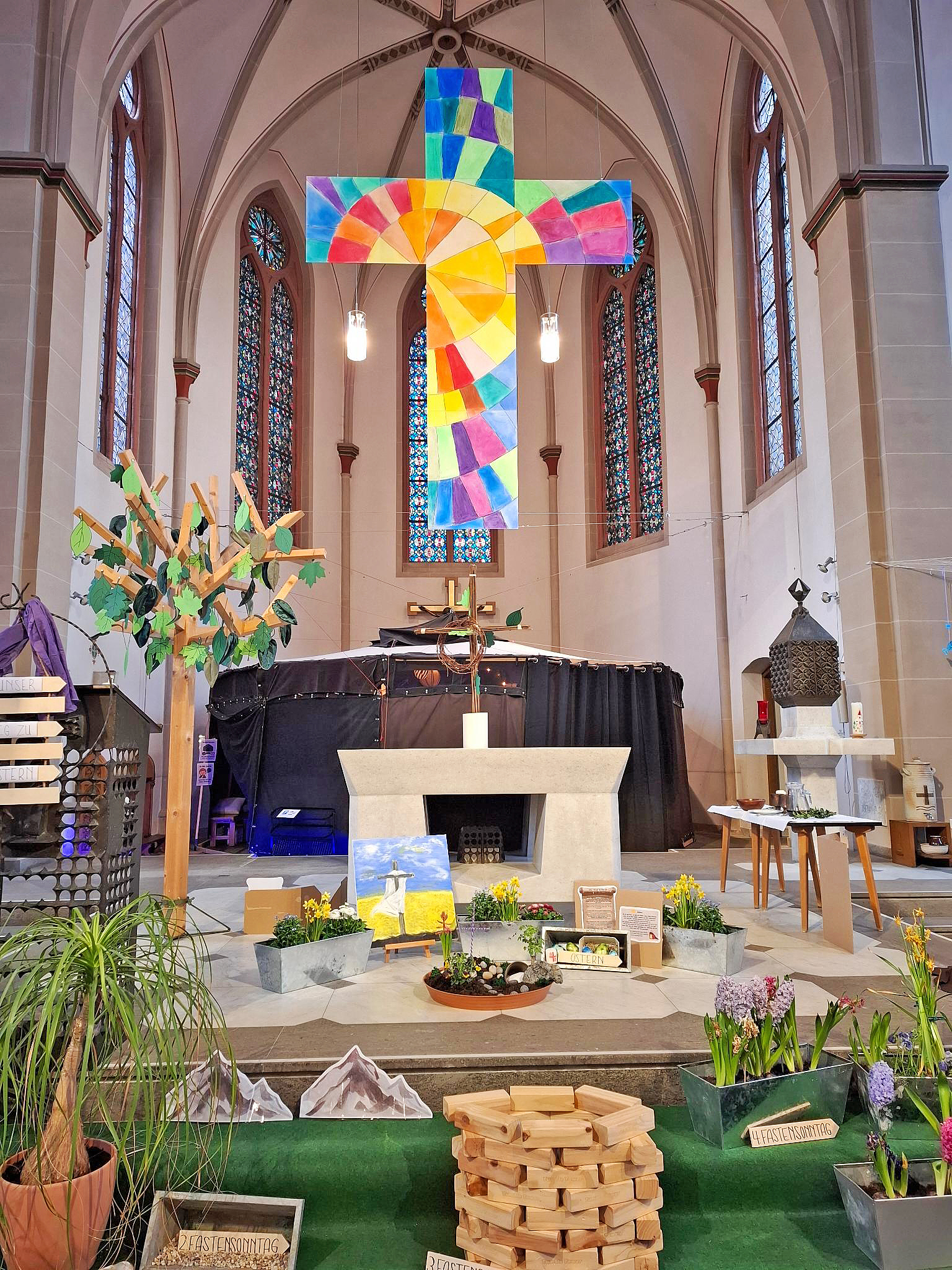 Blick auf Altar, hängendes buntes Kreuz, gebastelte Gegestände und Blumen vor dem Altar