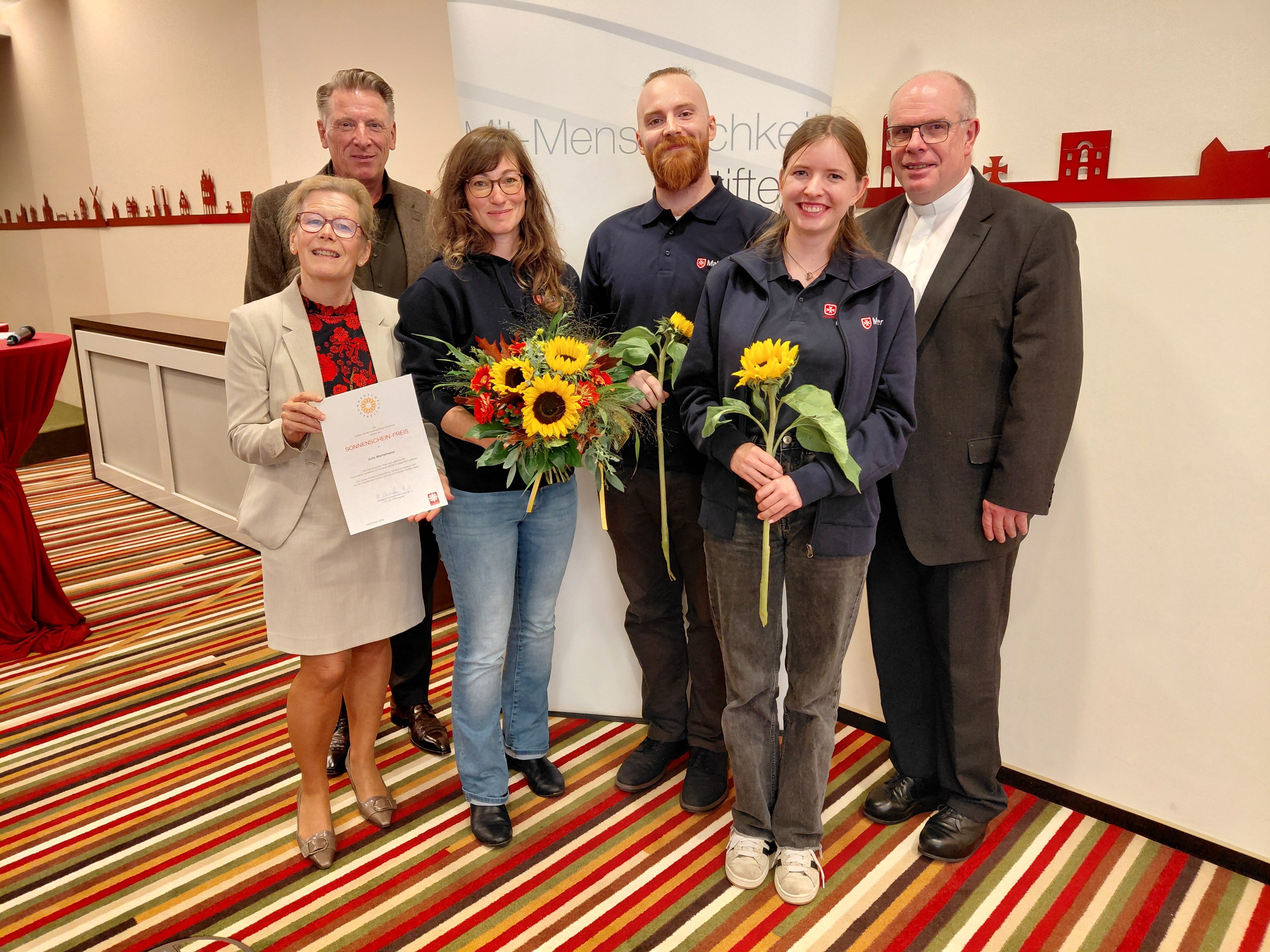 Fünf ältere Menschen und eine junge Frau vor einer Caritas-Fotowand mit einer Sonnenblume