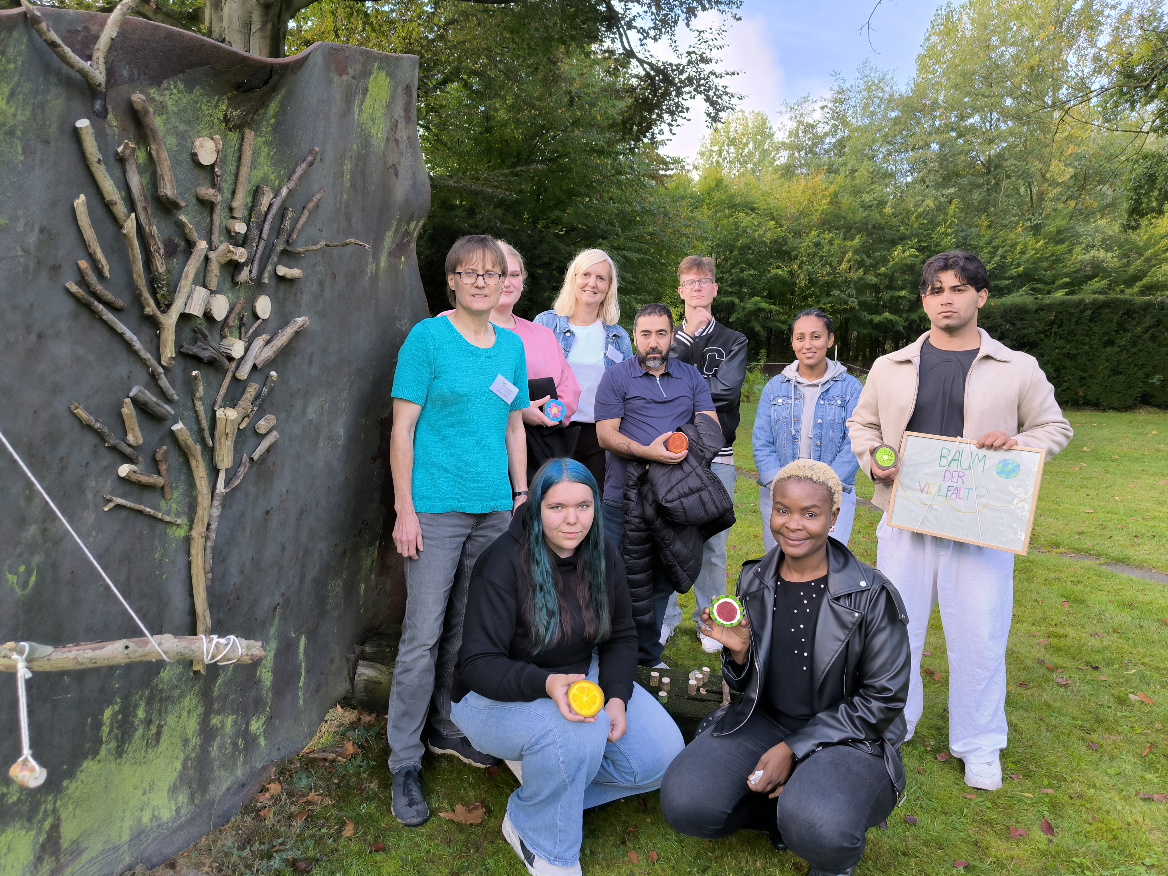 Eine Schulklasse und ihre Lehrerinnen stehen neben einer Metallwand, auf der ein Baum aus Holzstücken befestigt wurde. Ein Schüler trägt ein Schild mit der Aufschrift "Baum der Vielfalt" 
