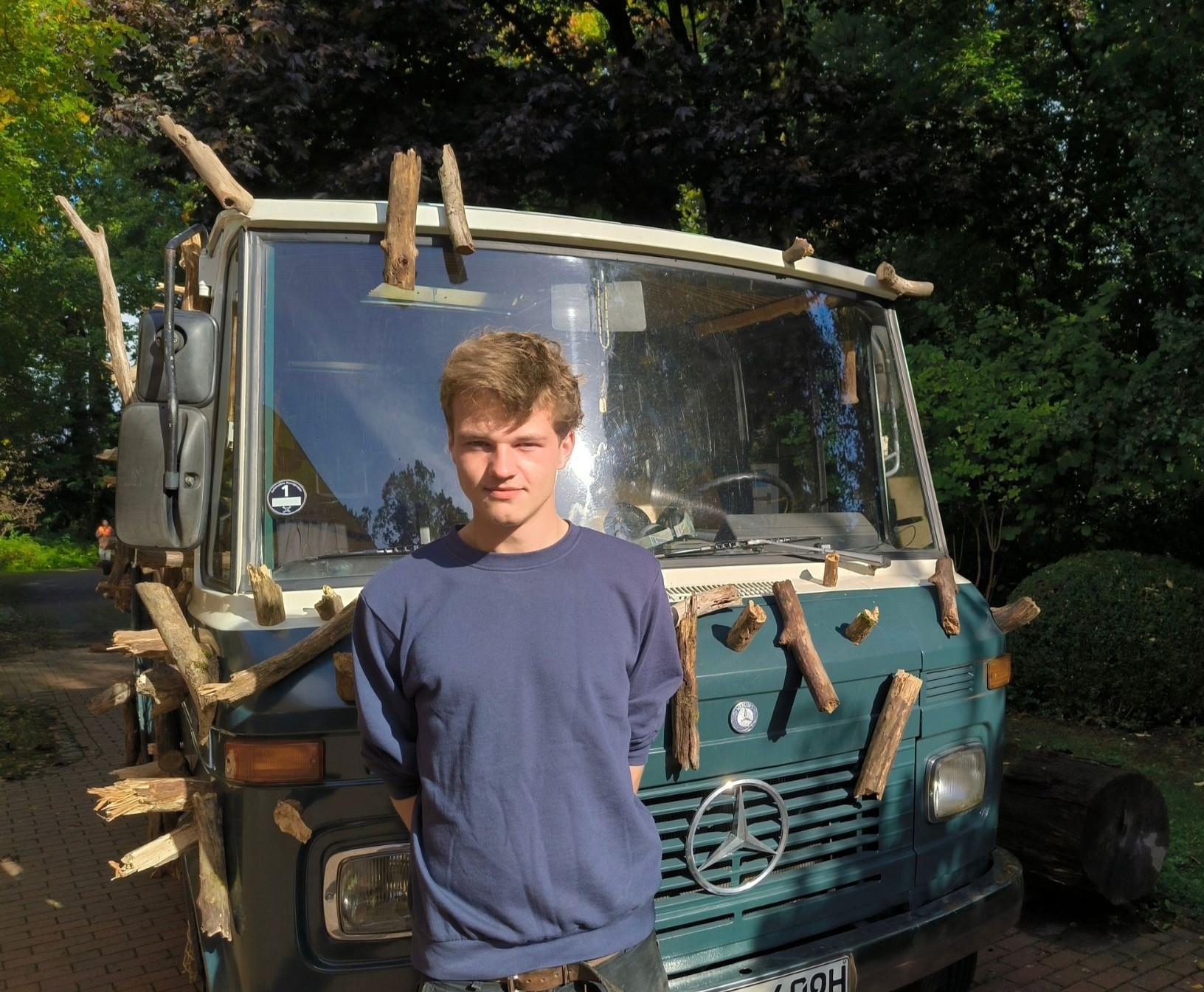 Ein junger Mann steht vor einem alten Mercedes-Kleinbus , der mit Holzstücken beklebt wurde