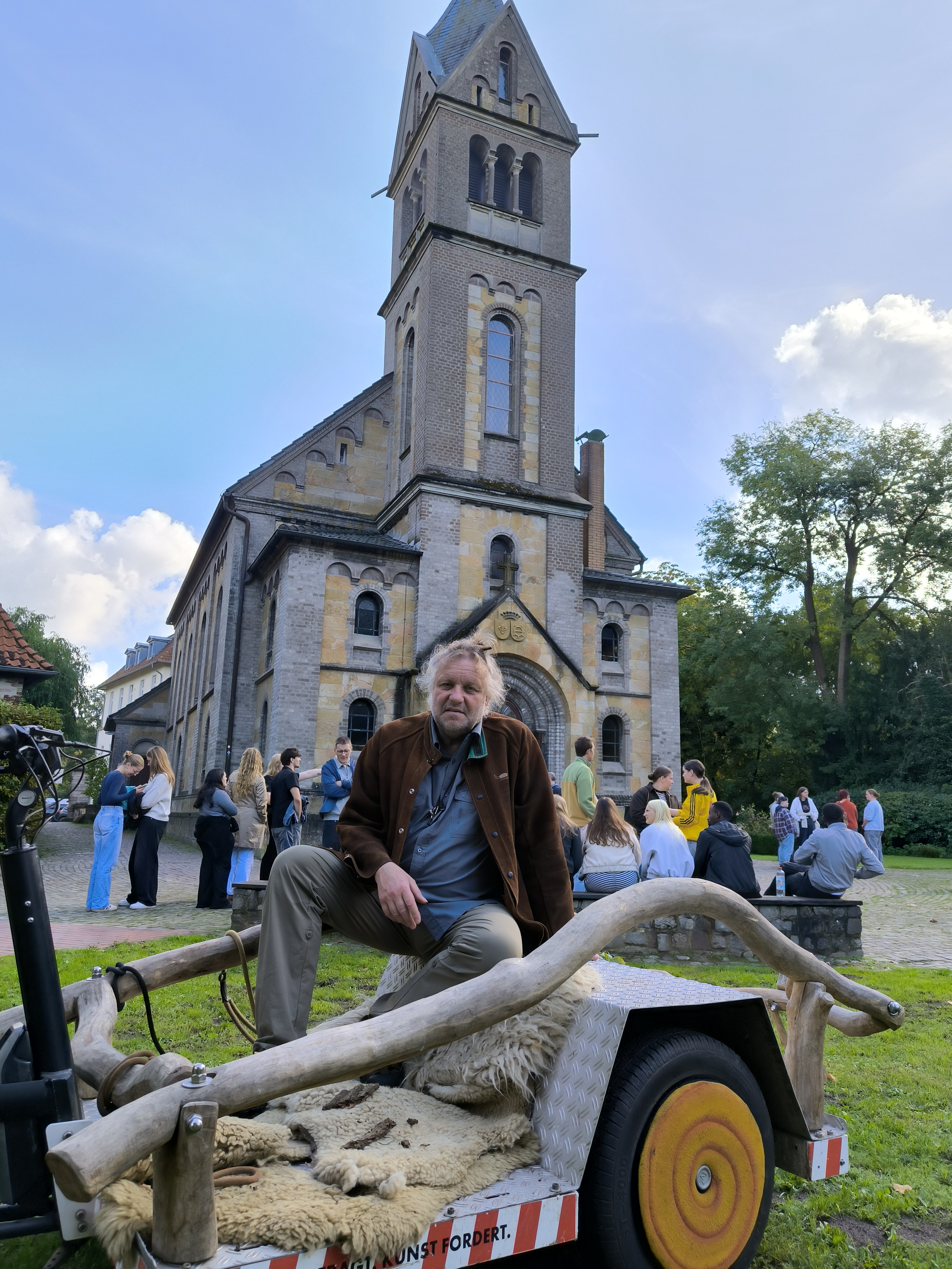 Der Künstler Volker-Johannes Trieb sitzt auf einem selbstgebauten Fahrradanhänger  und schaut in die Kamera. Im Hintergrund ist eine Kirche zu sehen. 