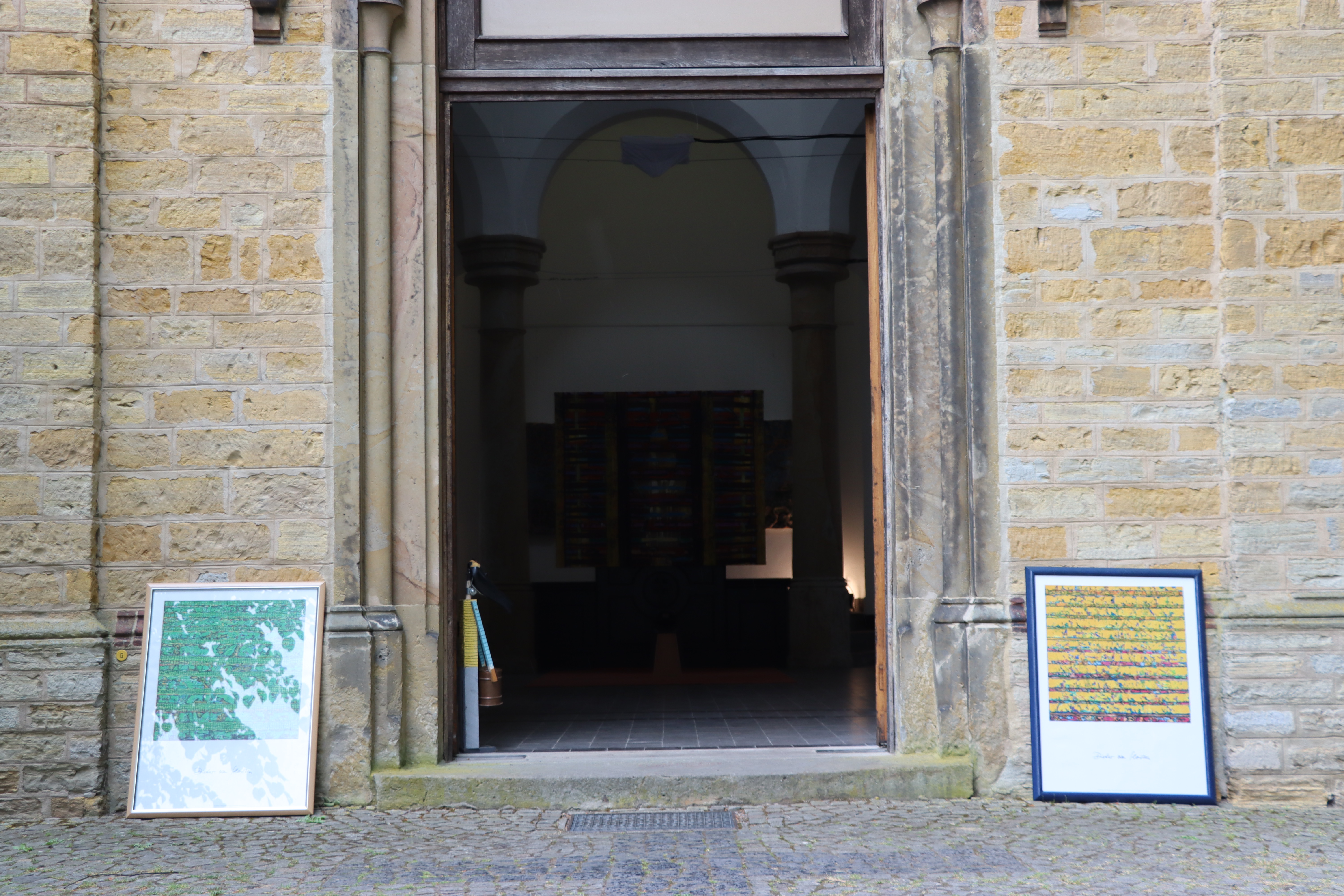 Kunst auf dem Osnabrücker Hasefriedhof. Eine alte Kapelle dient als Museum. Auf dem Bild sieht man den dunklen Eingang der Kapelle, die Tür ist geöffnet. Links und rechts lehnen Bilder am Boden des Eingangs. 