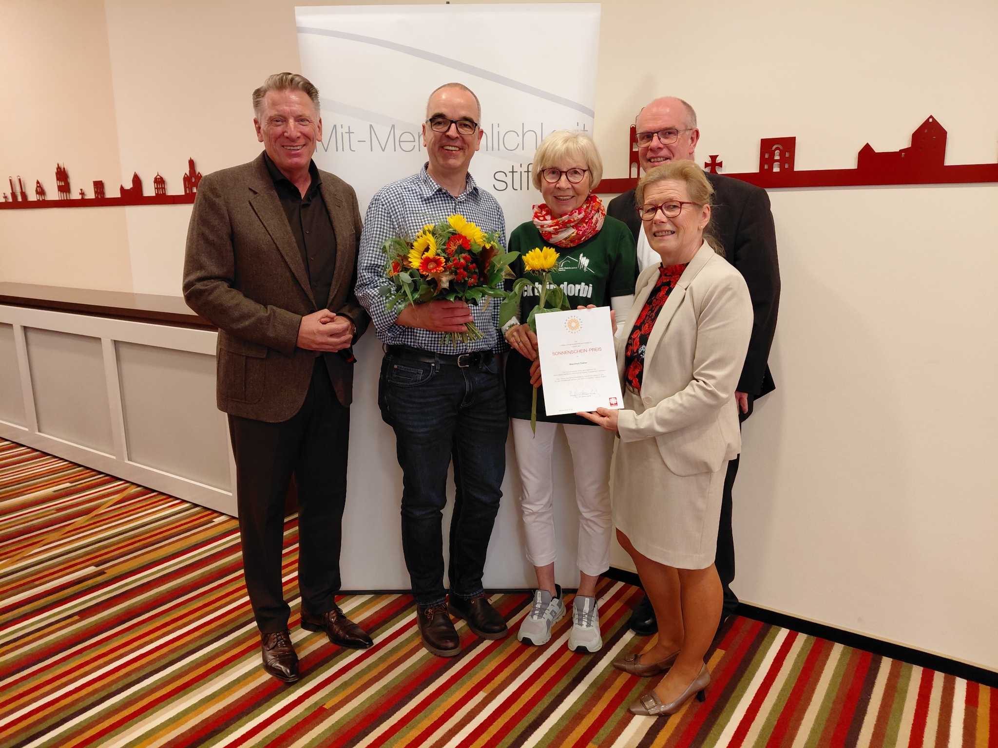 Fünf ältere Menschen vor einer Caritas-Fotowand mit einer Sonnenblume
