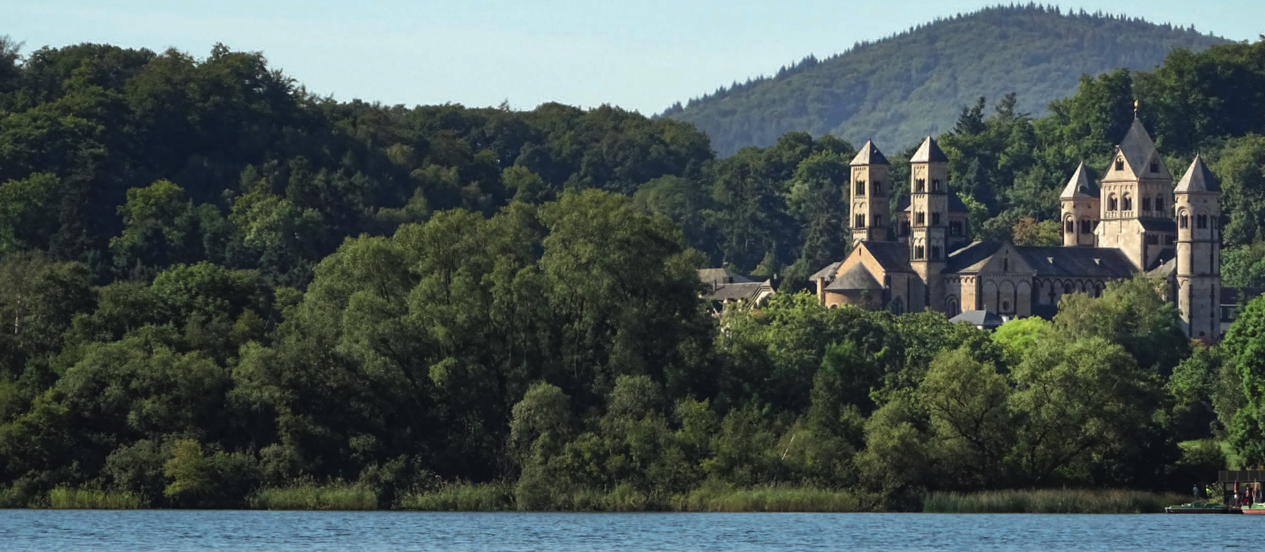Im Vordergrund ein See, im Hintergrund ein Gebäude mit Wald