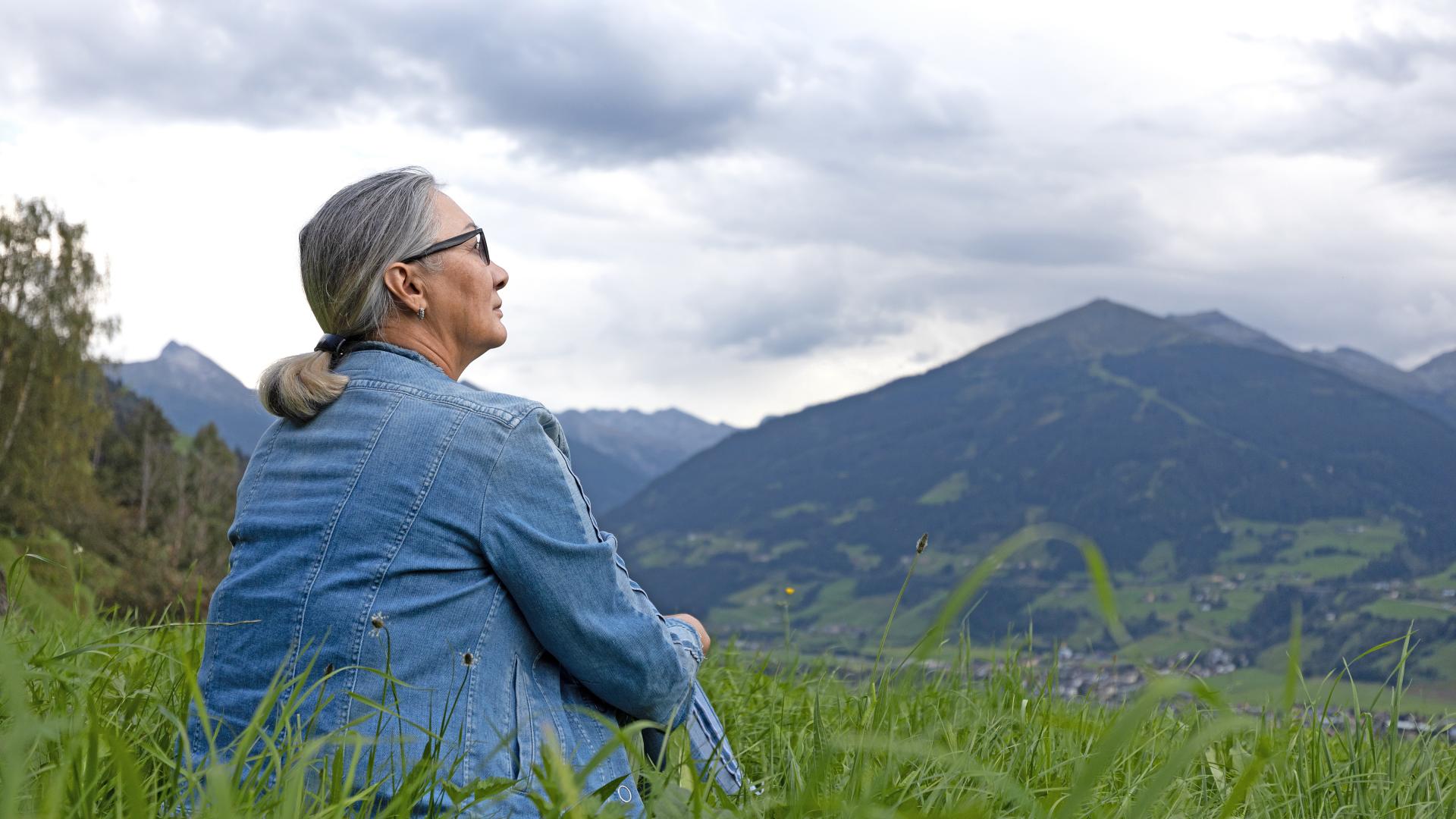 Eine grauhaarige Frau mit Pferdeschwanzfrisur und in Jeansjacke sitzt auf einer Wiese und betrachtet das Bergpanorama bei Salzburg. 