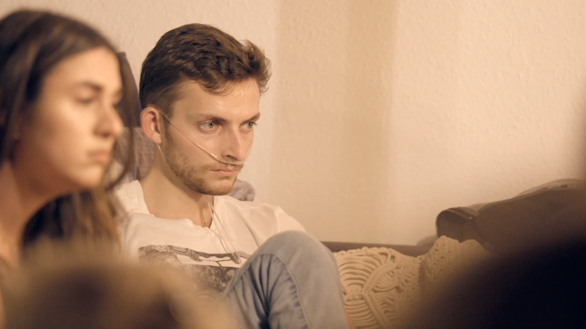 Philipp Mickenberger sitzt auf einer Couch mit Freunden, starrt ausdruckslos vor sich und erhält über einen dünnen Schlauch Sauerstoff