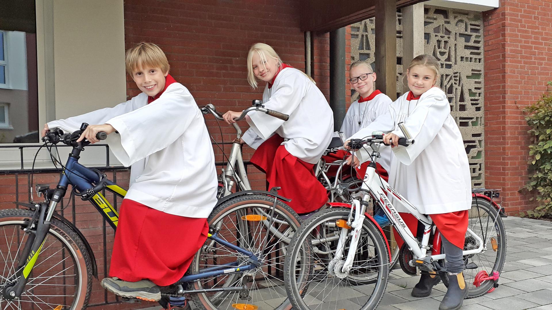 Vier Kinder sitzen in Ministrantengewändern auf Fahrrädern