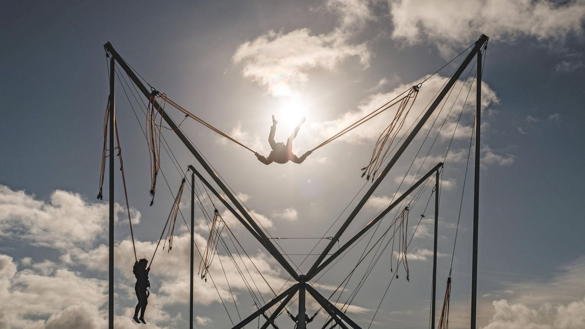 Zwei Silhouetten springen auf einem Trampolin mit Gummibefestigung in den Himmel. Die Sonne scheint im Hintergrund