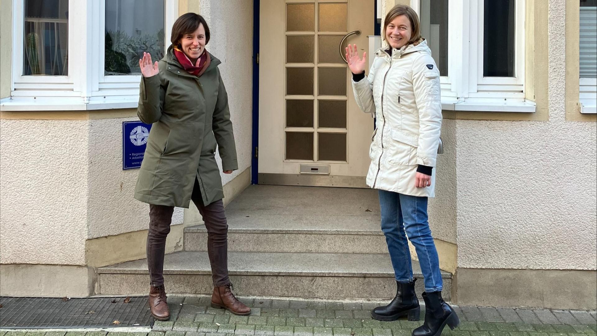 Zwei Frauen stehen winkend vor einem Gebäude