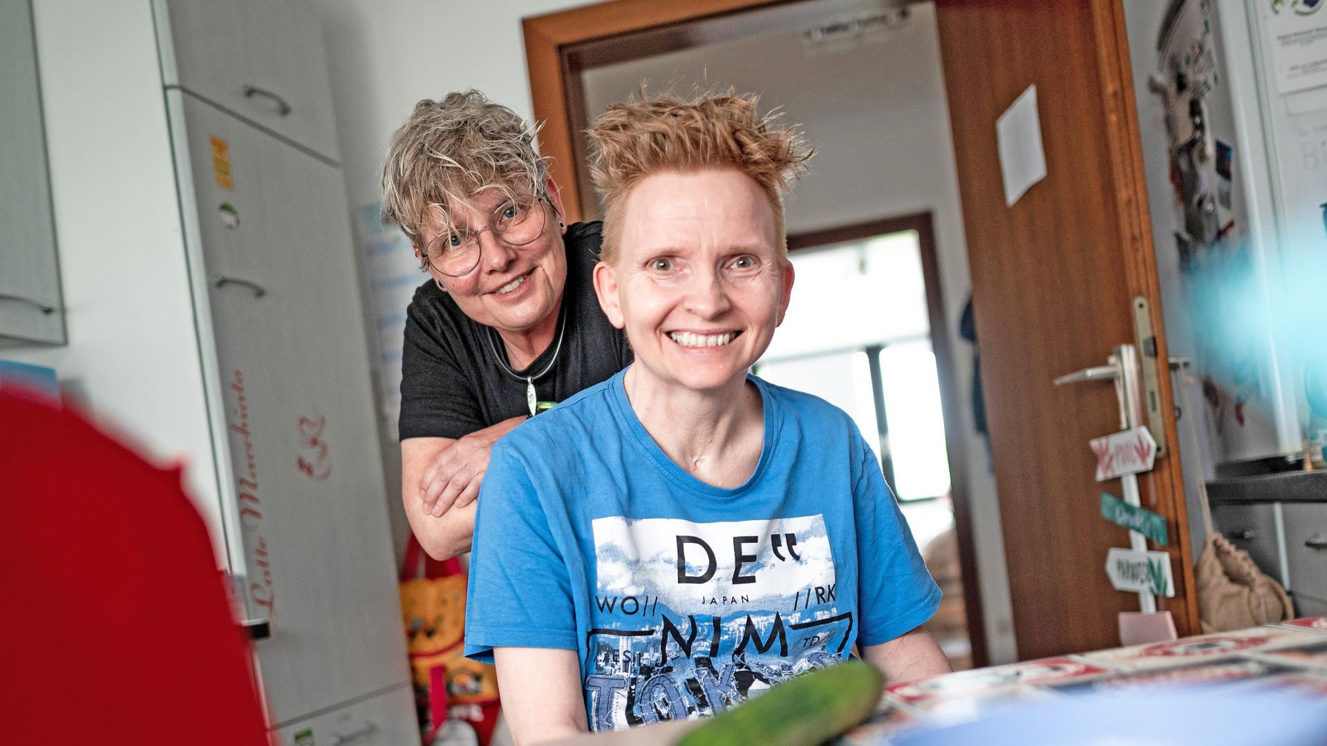 Eine Frau steht hinter einer anderen Frau, die am Küchentisch sitzt. Beide schauen in die Kamera.