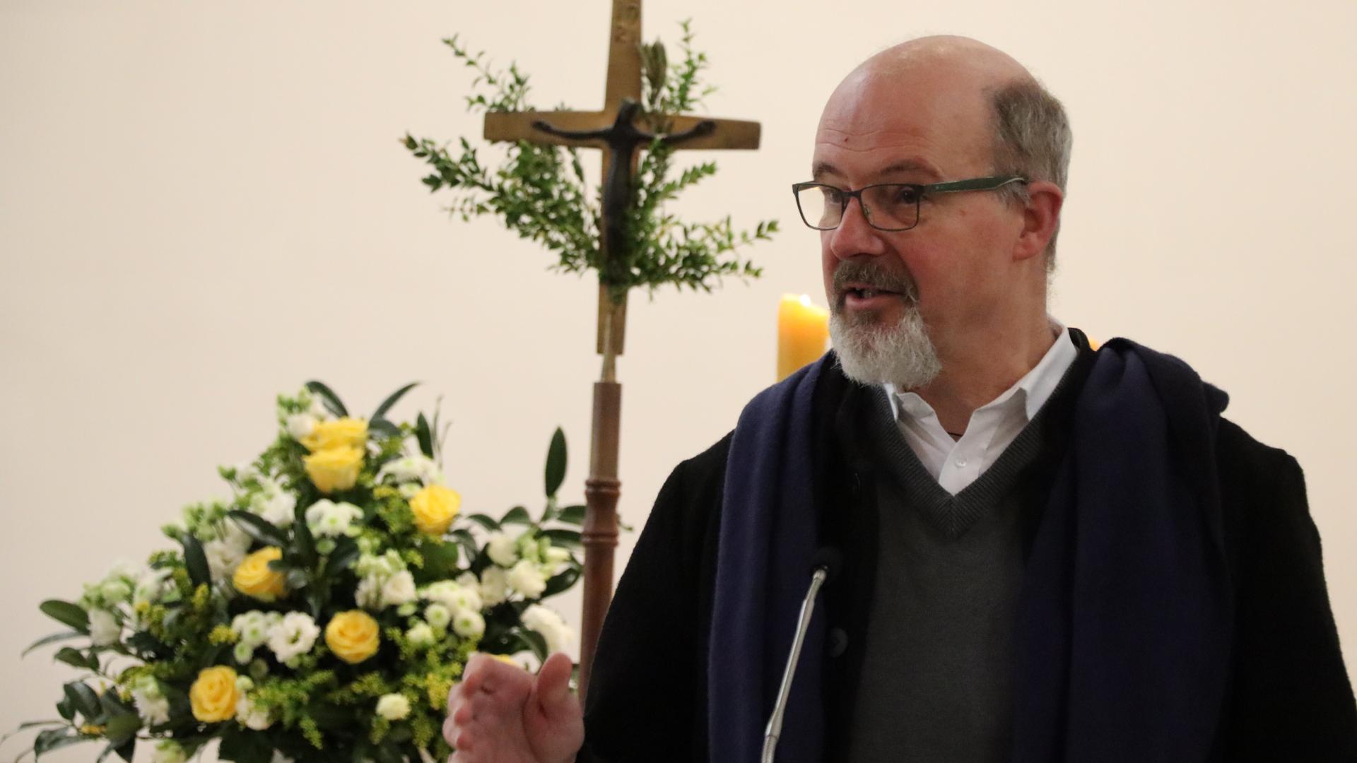 Ein Mann hält einen Vortrag, im Hintergrund sind ein Kreuz und Blumen