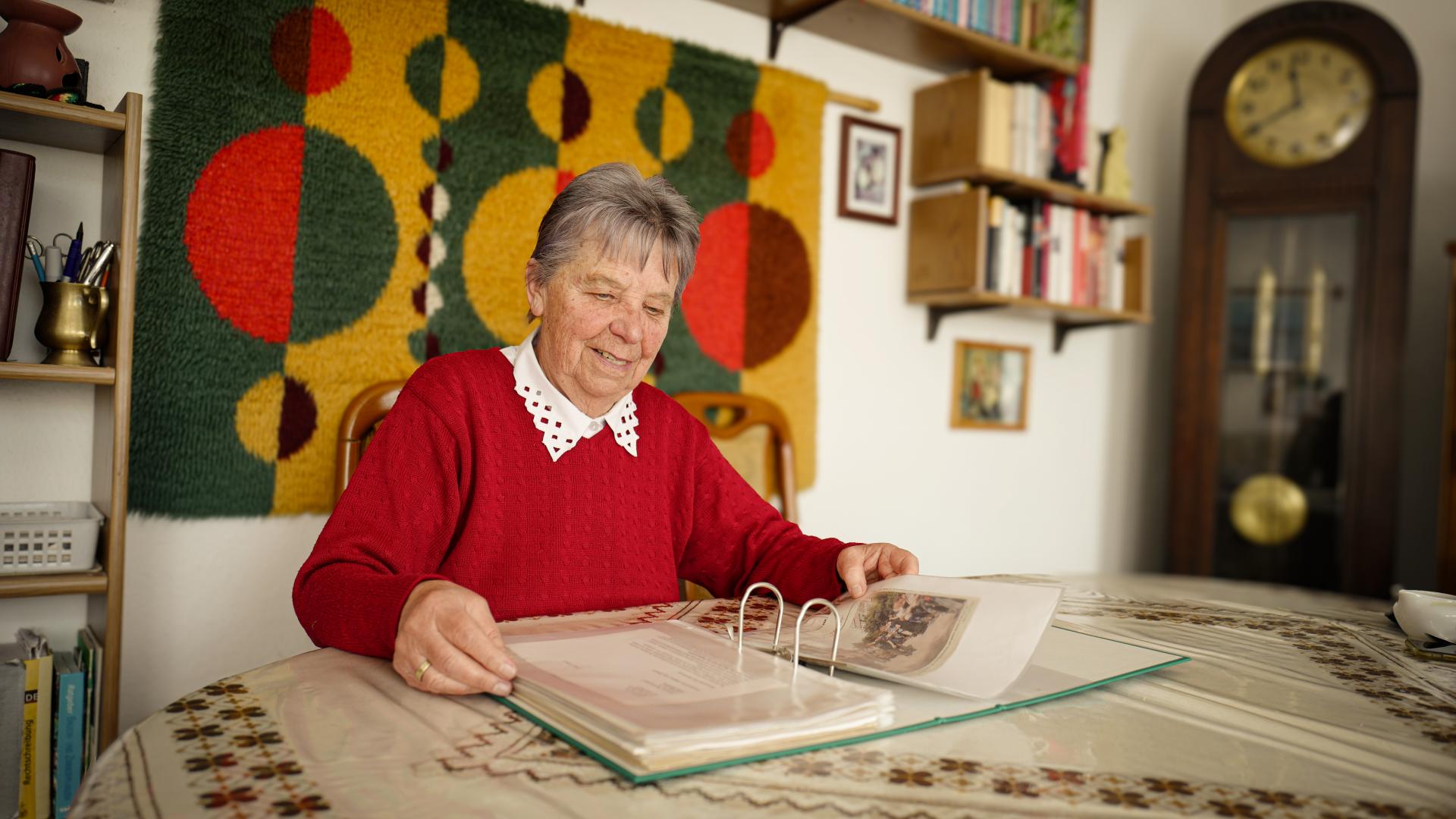 Eine Frau mit grauen Haaren sitzt in einem roten Pullover vor einem Tisch auf dem ein Aktenordner aufgeschlagen liegt