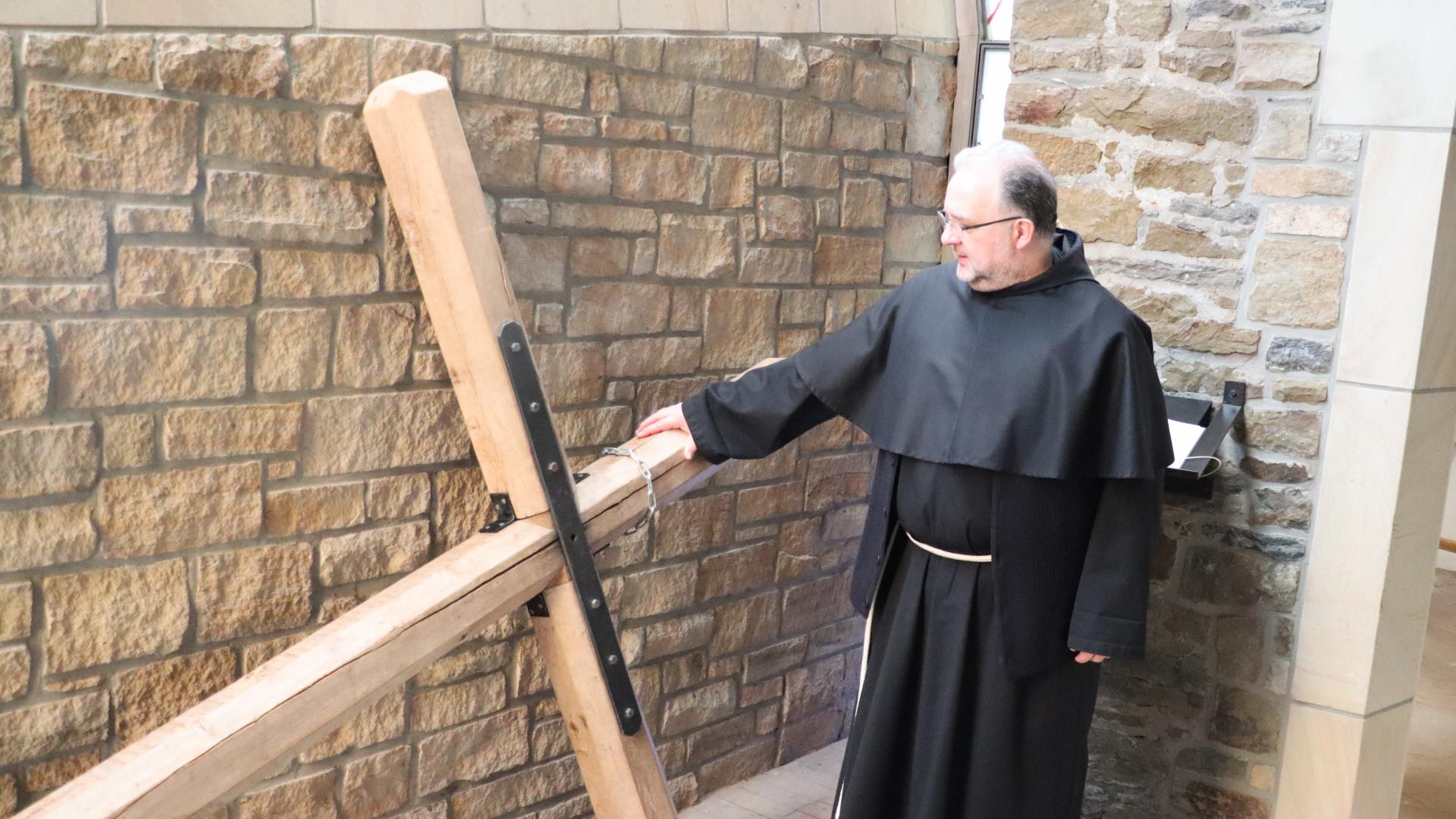 Ein Franziskanerpater steht neben einem schlichten Holzkreuz, das etwa drei Meter groß ist.