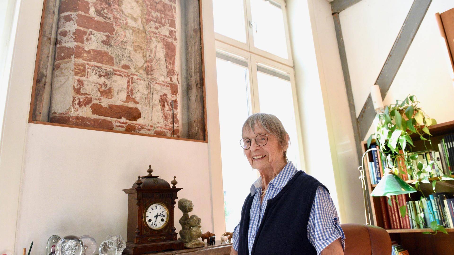 Eine Frau präsentiert Malereien in ihrer Wohnung und lächelt in die Kamera.