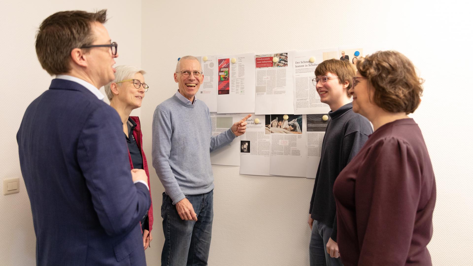 An einer Wand hängen mehrere Zeitungsseiten. Davor stehen fünf Frauen und Männer.