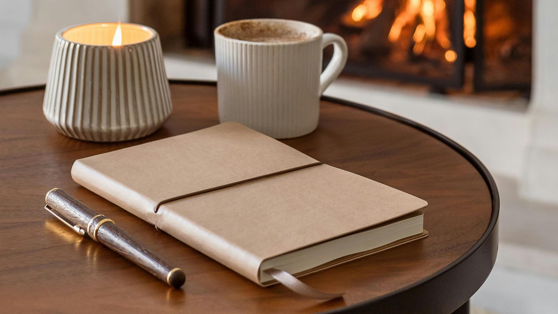 Kerze, Kafeetasse, Buch und Stift liegen auf einem Holztisch, Im Hintergrund ein Kamin mit Feuer