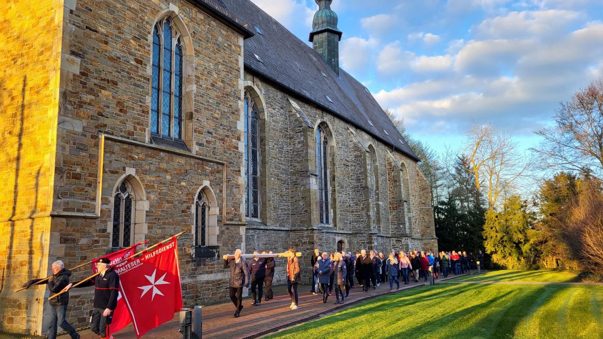 Eine Menschengruppe zieht hinter einem Banner des Malteserordens um die Kirche in Lage