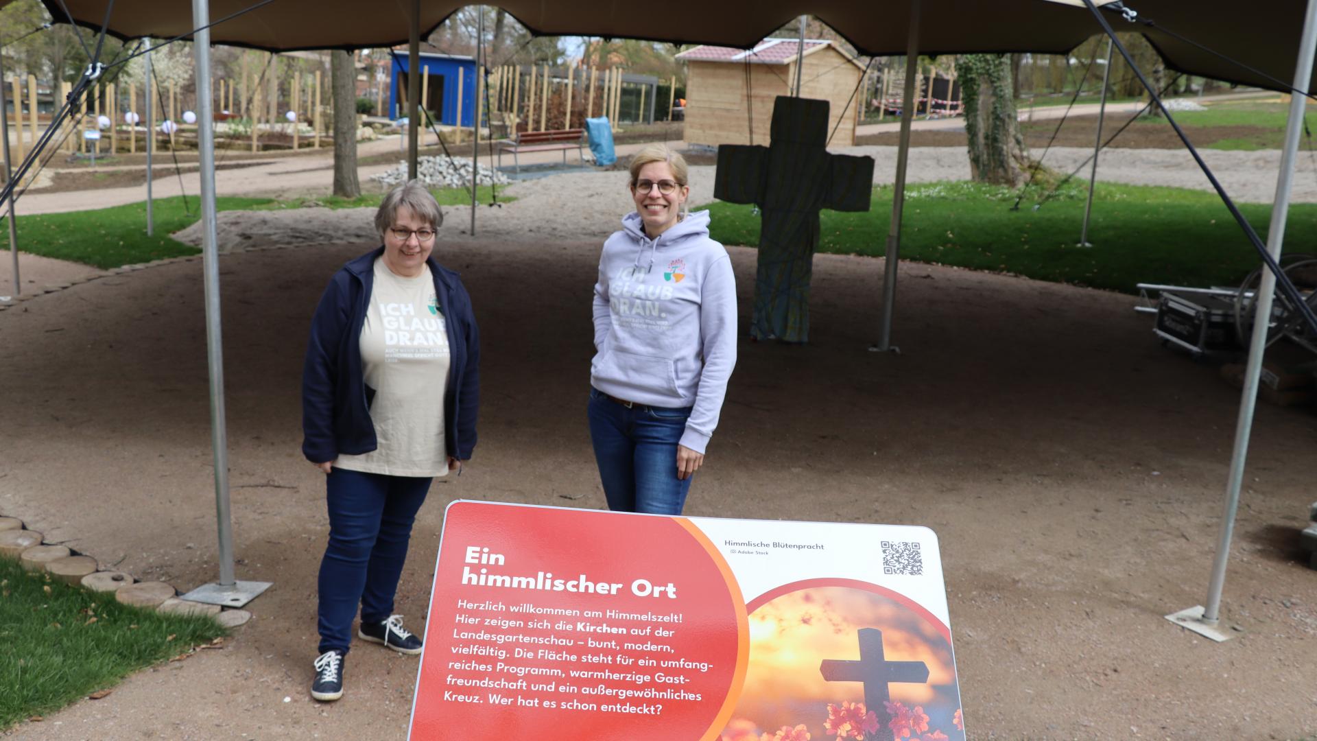 Zwei Fauen stehen vor einemZelt, vor ihnen steht ein Schild mit der Überschrift "Ein himmlischer Ort"