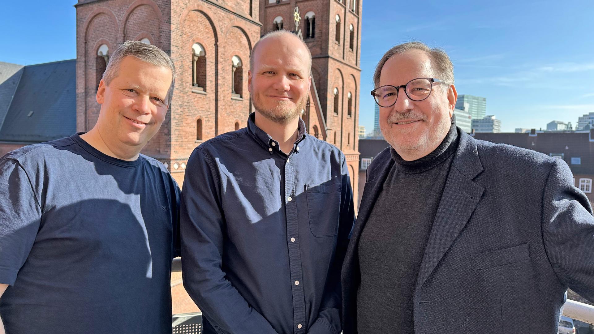 Drei Herren lächeln in die Kamera, hinter ihnen ist der St. Marien-Dom zu sehen.