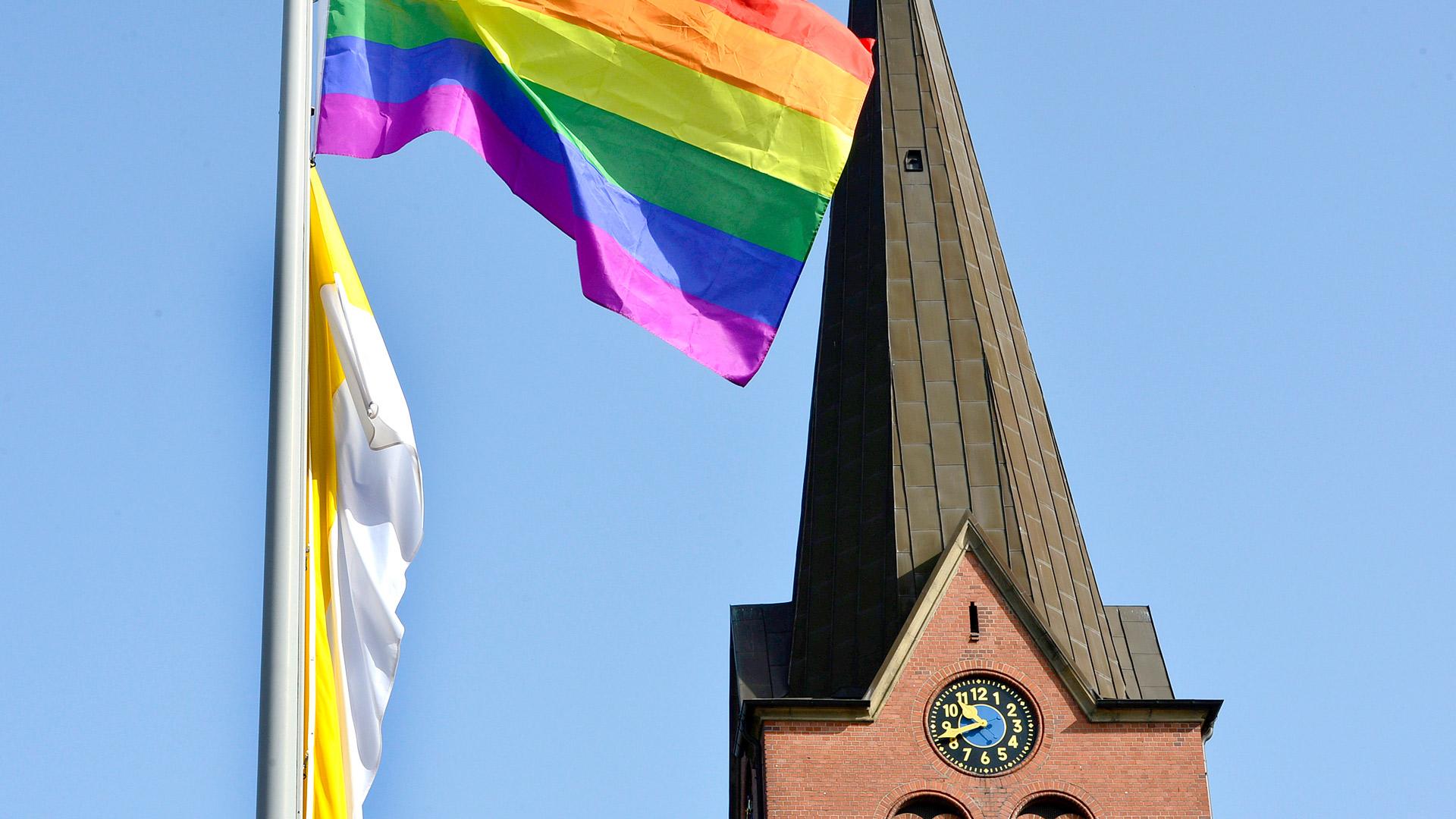 eine Regenbogenfahne weht vor einem Kirchturm