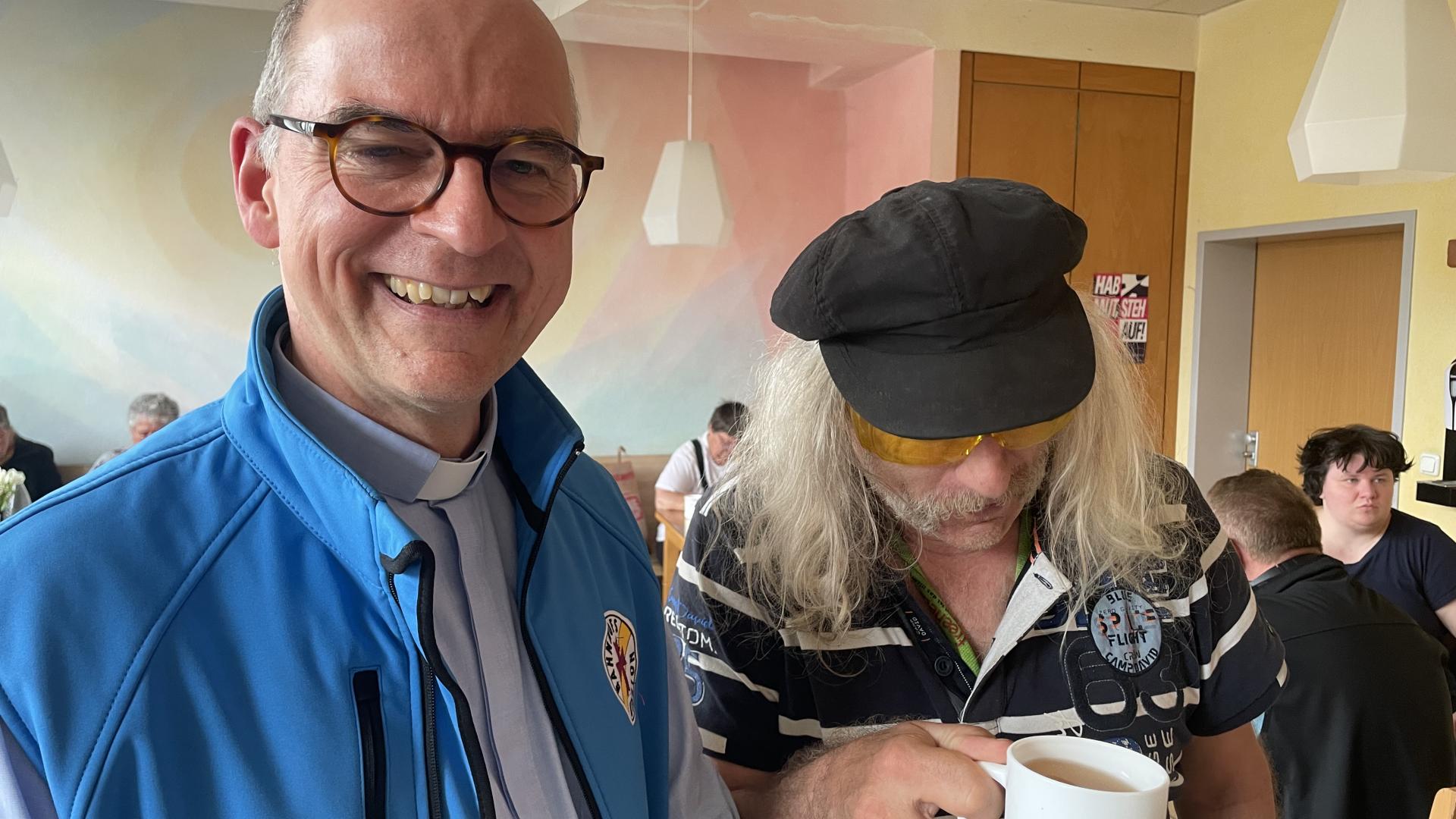 Bischof von Würzburg mit Frau in Bahnhofsmission. Sie hält eine Tasse, er trägt blaue Weste. 
