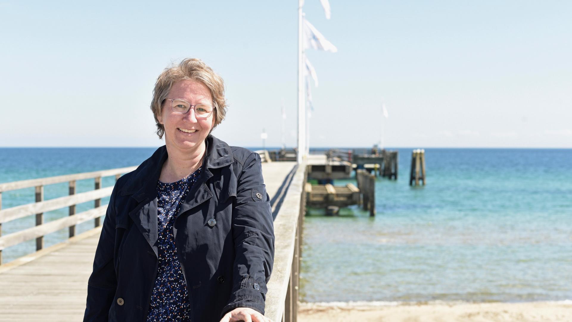 Eine Frau steht auf einer Seebrücke, im Hintergrund das Meer. Sie lächelt in die Kamera.