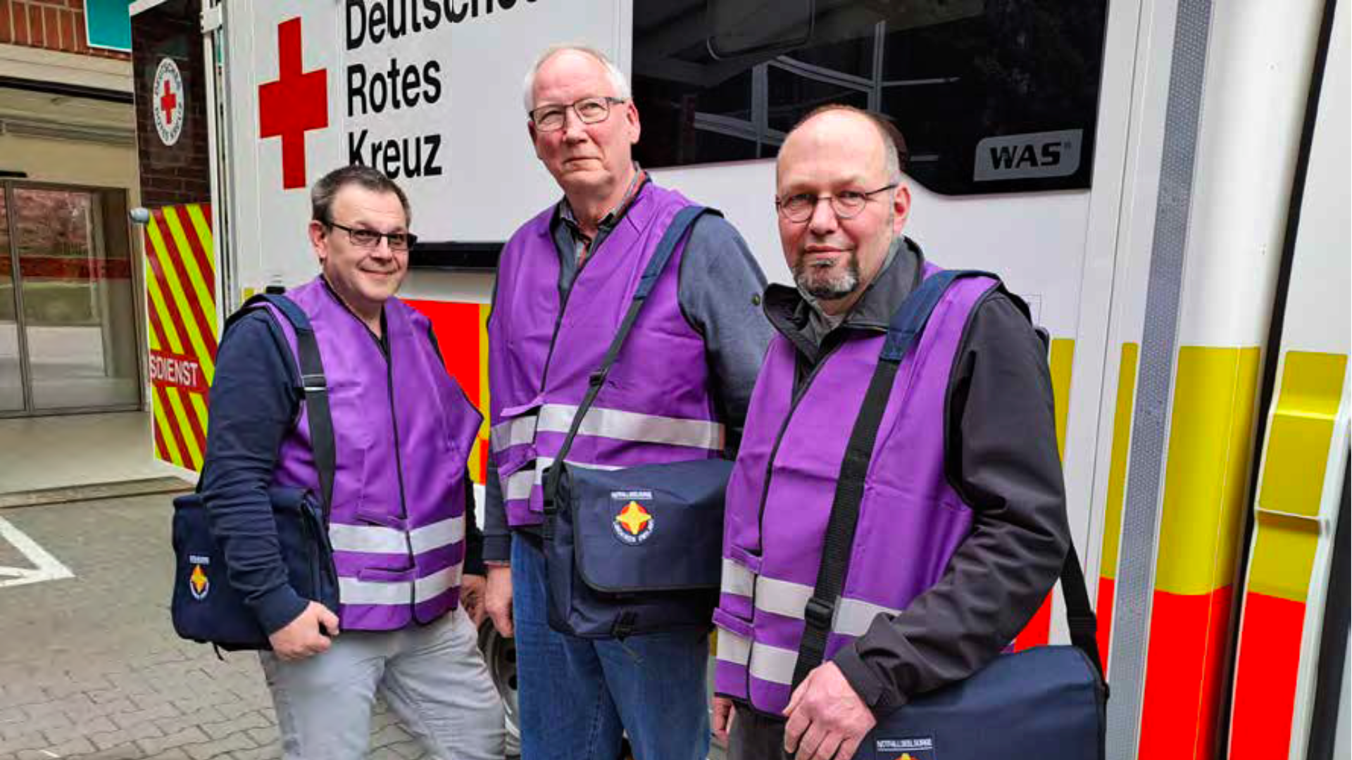 Drei Männer in lila Warnwesten vor einem Rettungswagen.