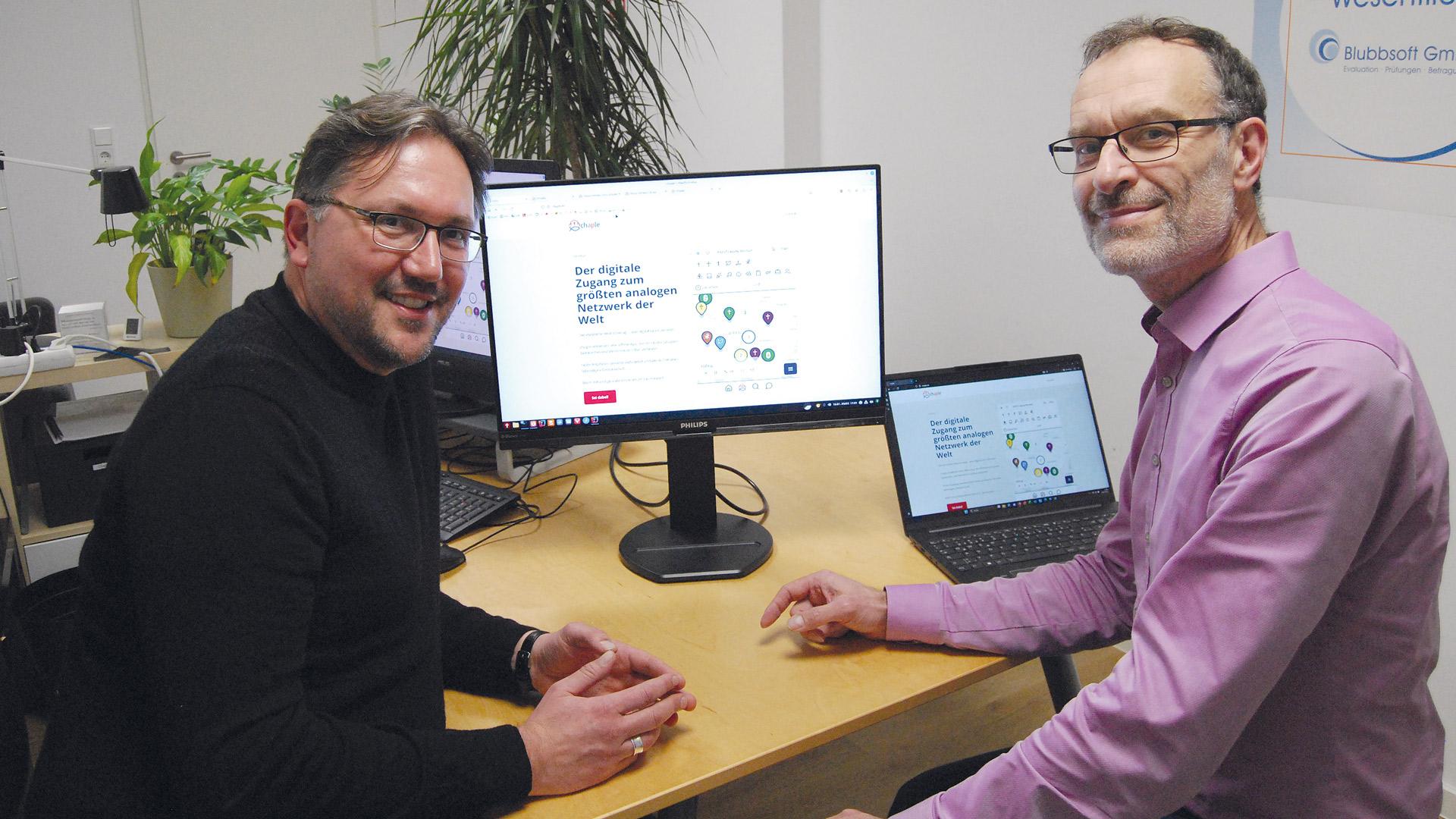 Lukasz Kopinke (links) und Frieder Nollau sitzen vor einem Schreibtisch, auf einem Computerbildschirm ist die Website chaple.de geöffnet