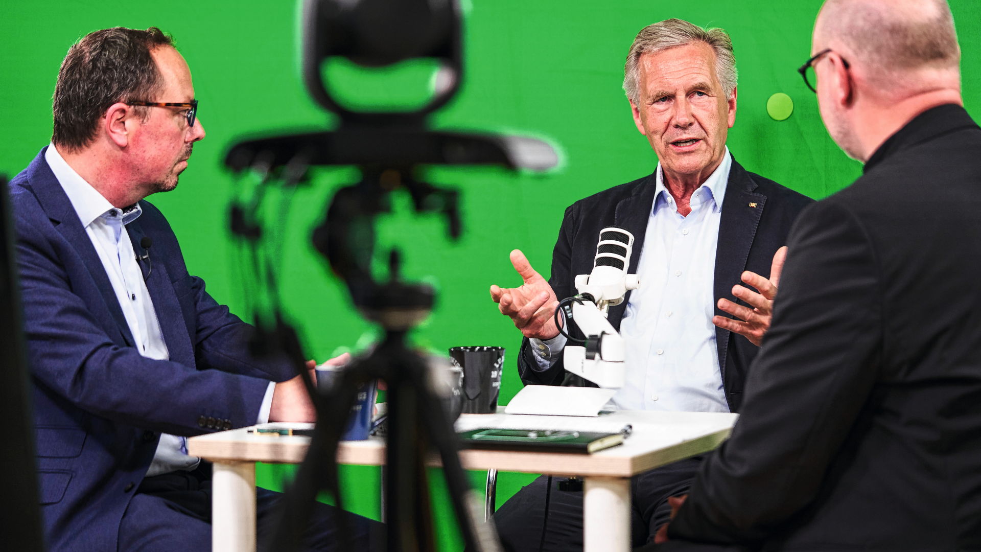 Drei Männer sitzen vor einem Greenscreen an Mikrofonen