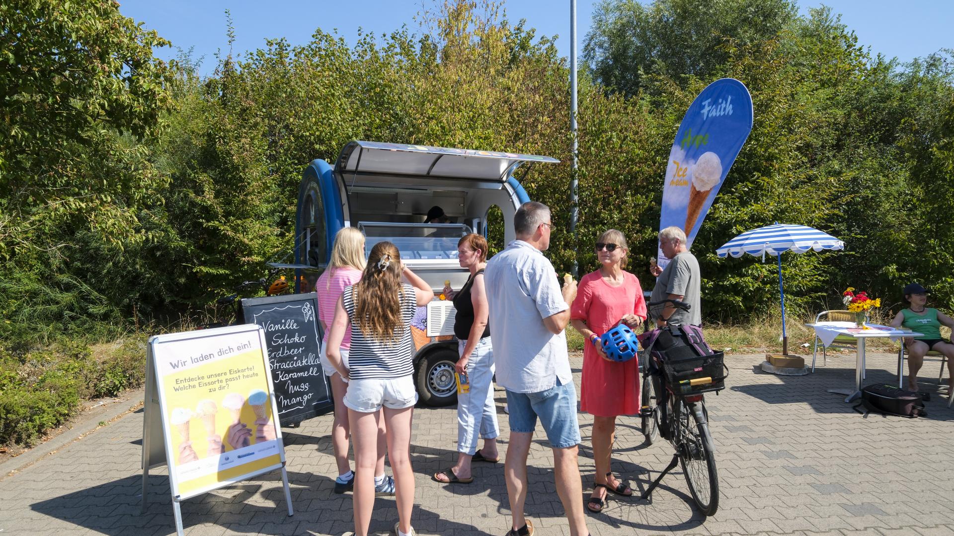 Ein Eiswagen steht auf einem Parkplatz, Menschen stehen drum herum in der Sonne