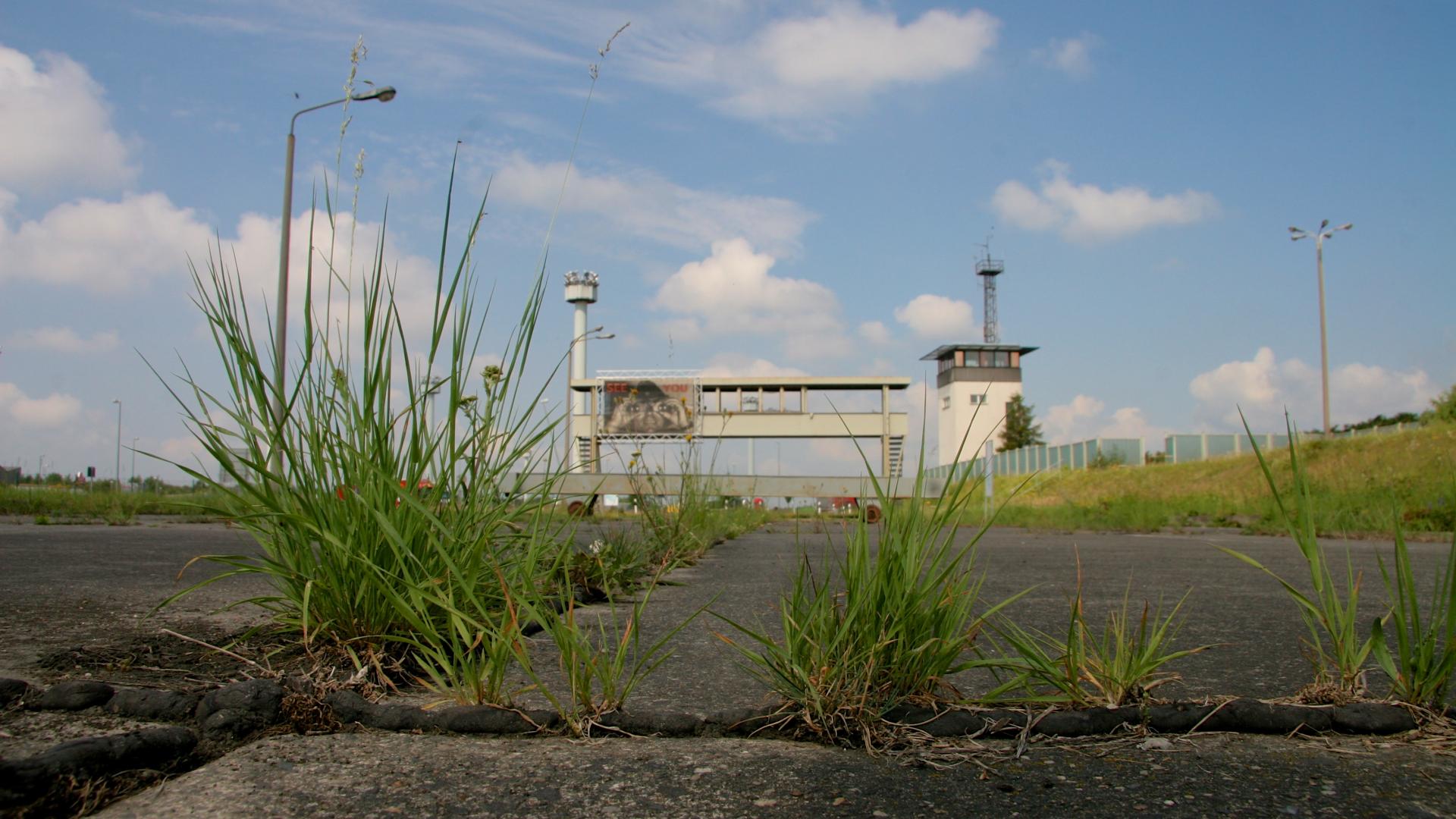 Im Vordergrund wächst Gras auf einer Betónautobahn, im Hintergrund sind Reste von Grezbefestigungen zu sehen