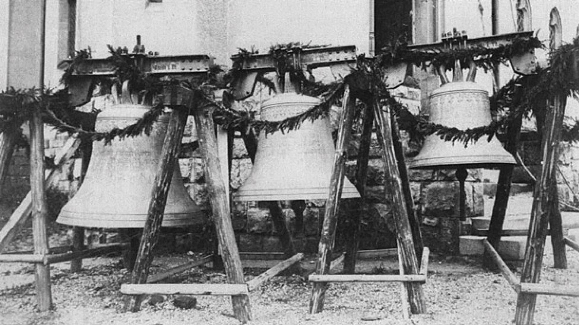 Schwarz-Weiß-Aufnahme von drei Glocken, die geschmückt vor einer Kirche stehen