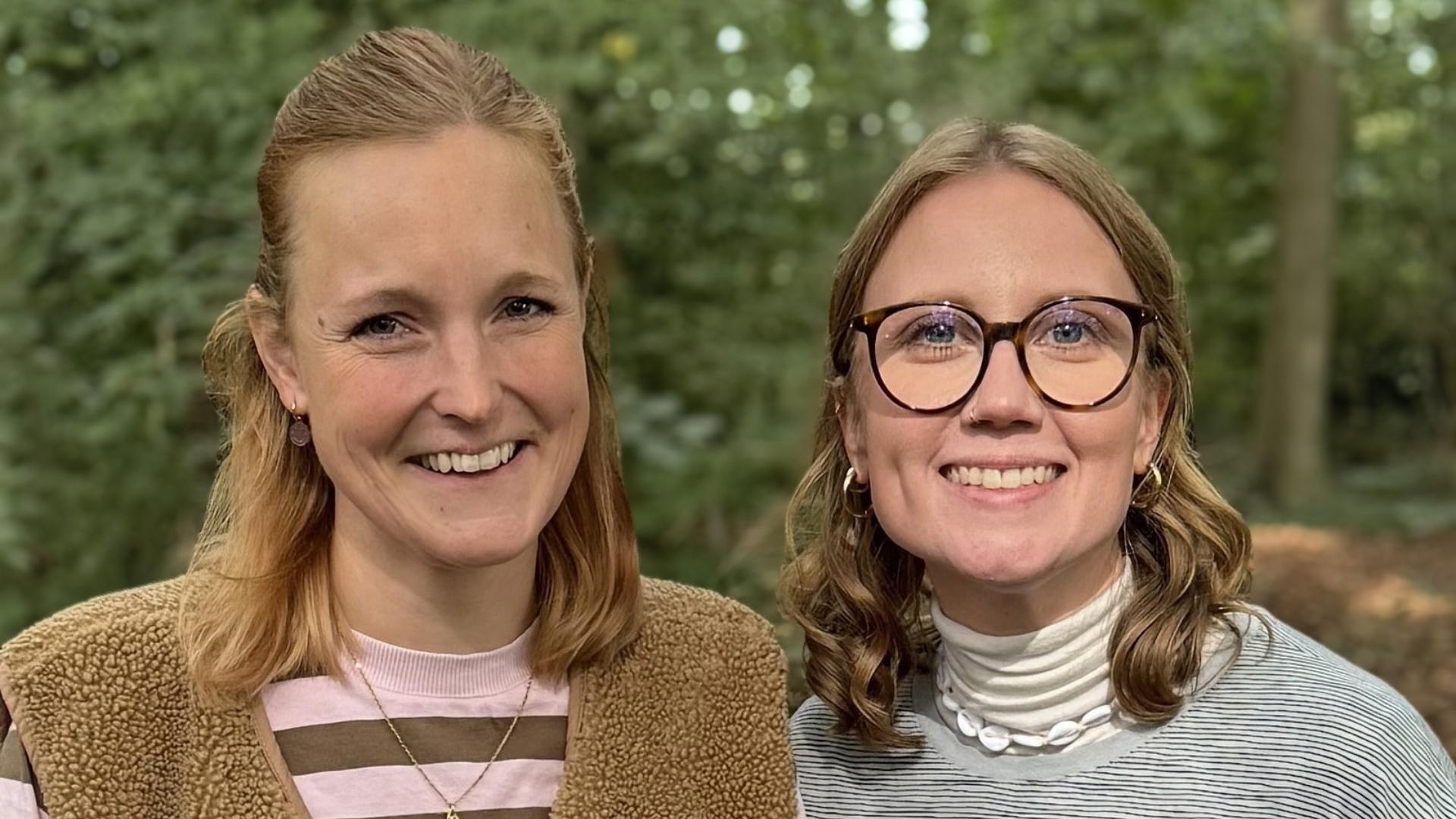 Zwei junge Frauen stehen vor Bäumen und lächeln in die Kamera