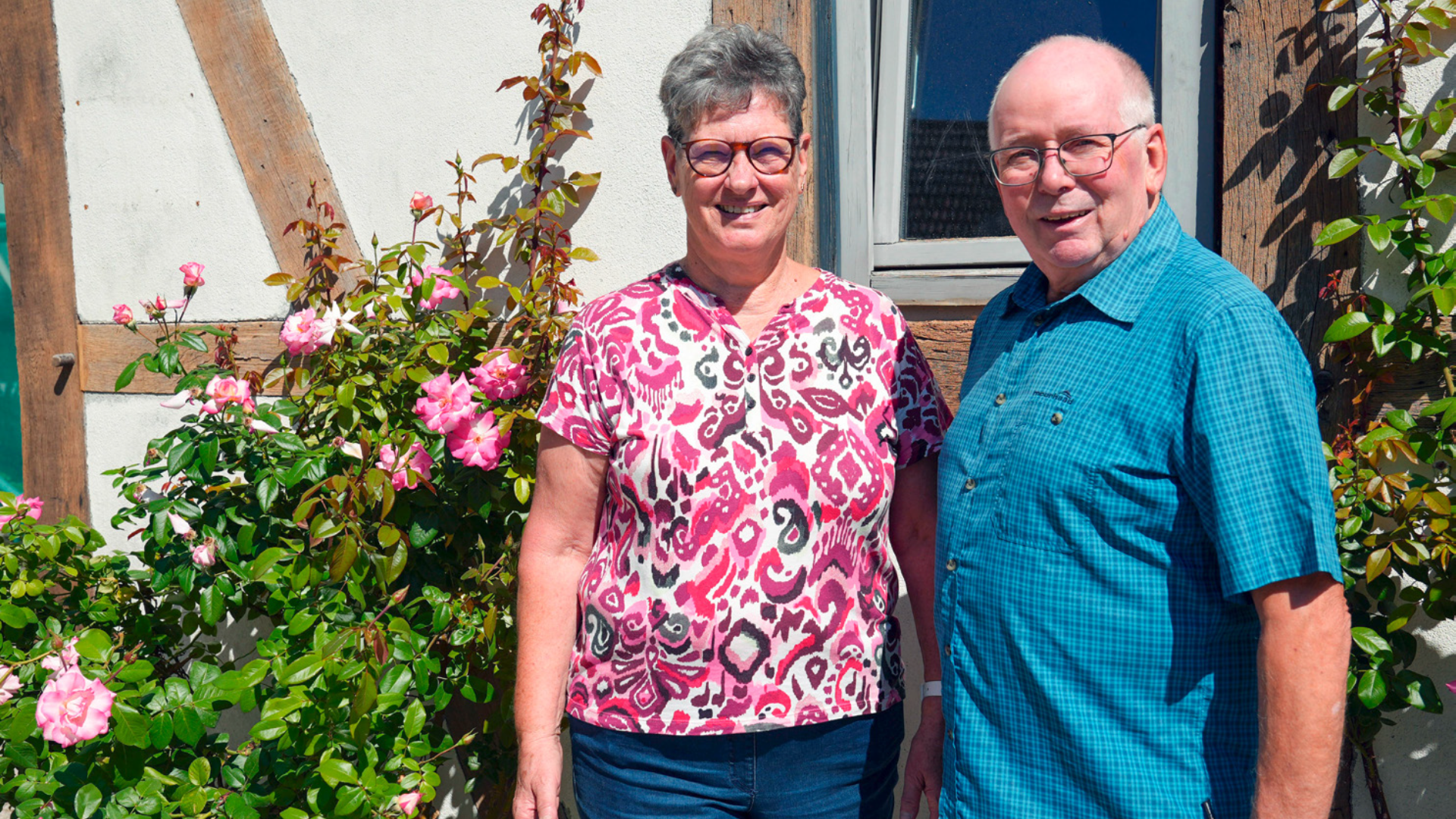 Zwei Menschen stehen vor einem Rosenstrauch und lächeln in die Kamera, ein Mann und eine Frau.