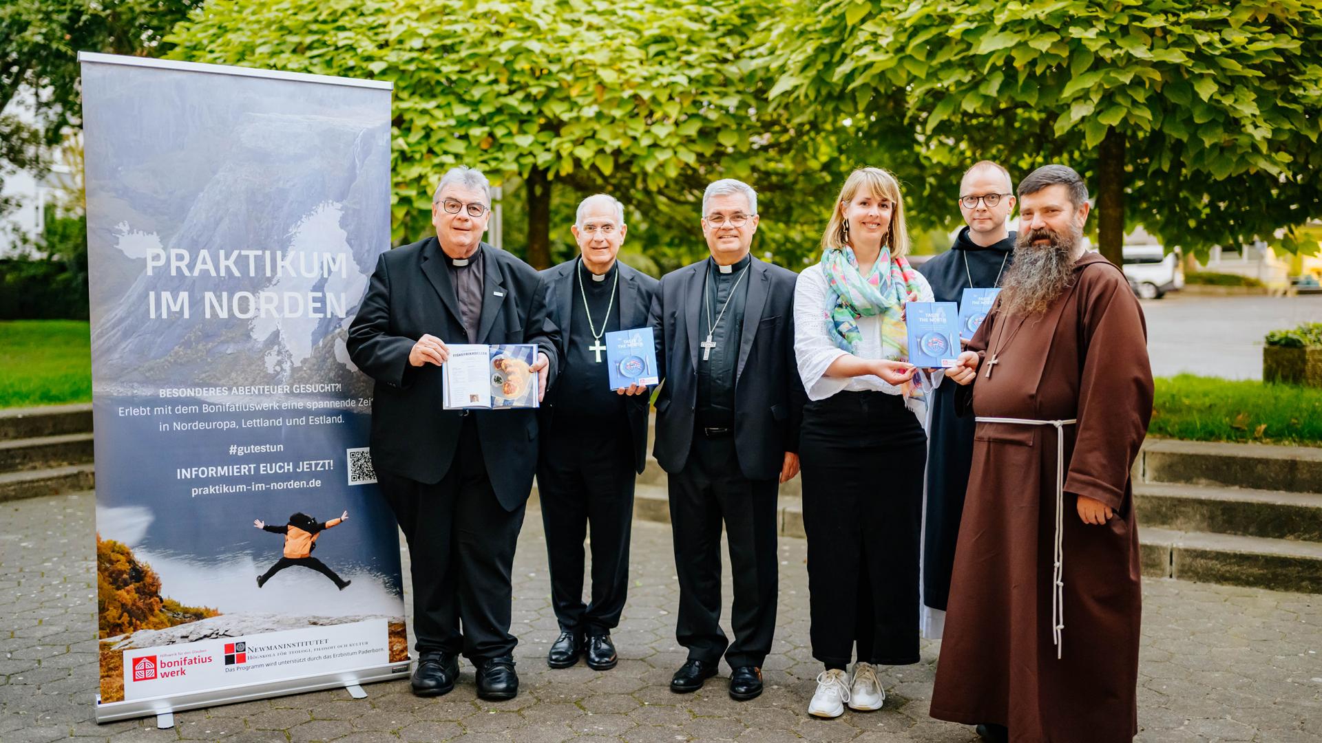 Vertreter und eine Vertreterin des Bonifatiuswerks, teilweise mit Mönchskutte bekleidet, stehen vor Bäumen und präsentieren das neue Kochbuch